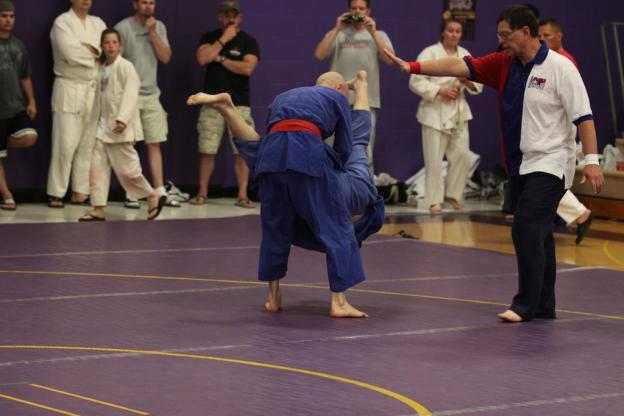 This photo shows a freestyle judo match on a circular mat and the referee calling matte and using a hand signal to have the timekeeper stop the clock.