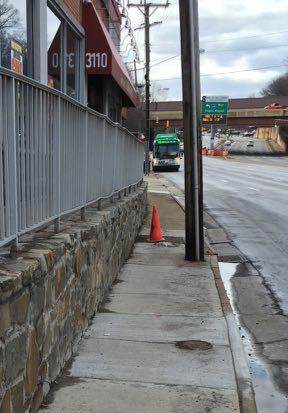 On the south side of the street, the sidewalk is generally in poor condition, with sections that are eroded (marked with an orange traffic cone), and low voltage power lines run the length of the