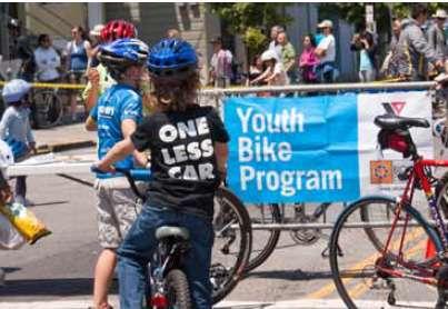 PAGE 57. Image: Child on bike wearing ONE LESS CAR shirt. Youth Bike Program banner in the background.