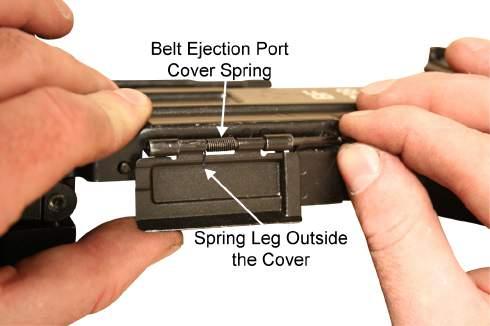 Align the belt ejection port cover spring with the pin, ensuring the left leg is outside the cover and the right leg is behind the cover.