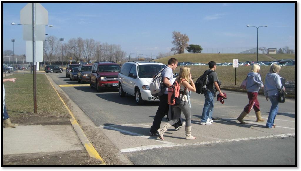Students crossing traffic flow entering and exiting the parking lot after dismissal. The crosswalk striping is not very visible and there are no pedestrian ramps for the crossing.